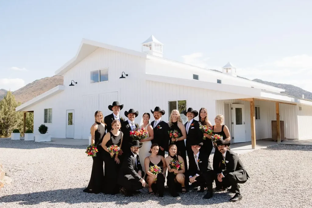 Group Wedding Portraits at White Barn Wedding Venue. Bride and groom surrounded by bridal party in matching sleek black wedding attire.
