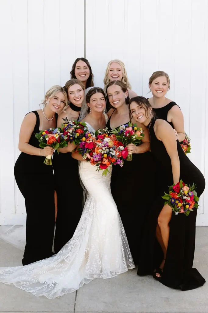 Sleek Black Bridesmaid Dresses with Bright Wildflower Bouquets. Bride standing with bridesmaids, bright florals in hand, at modern barn venue with white walls.