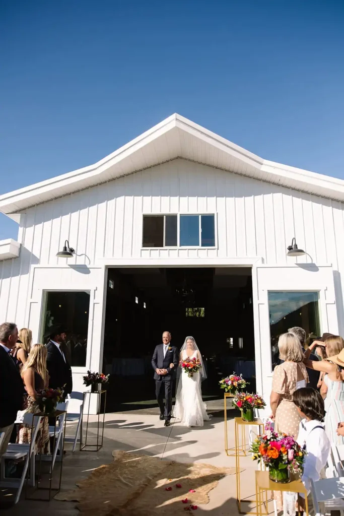 Bride walking down the aisle toward groom in rustic lakeside ceremony setup.