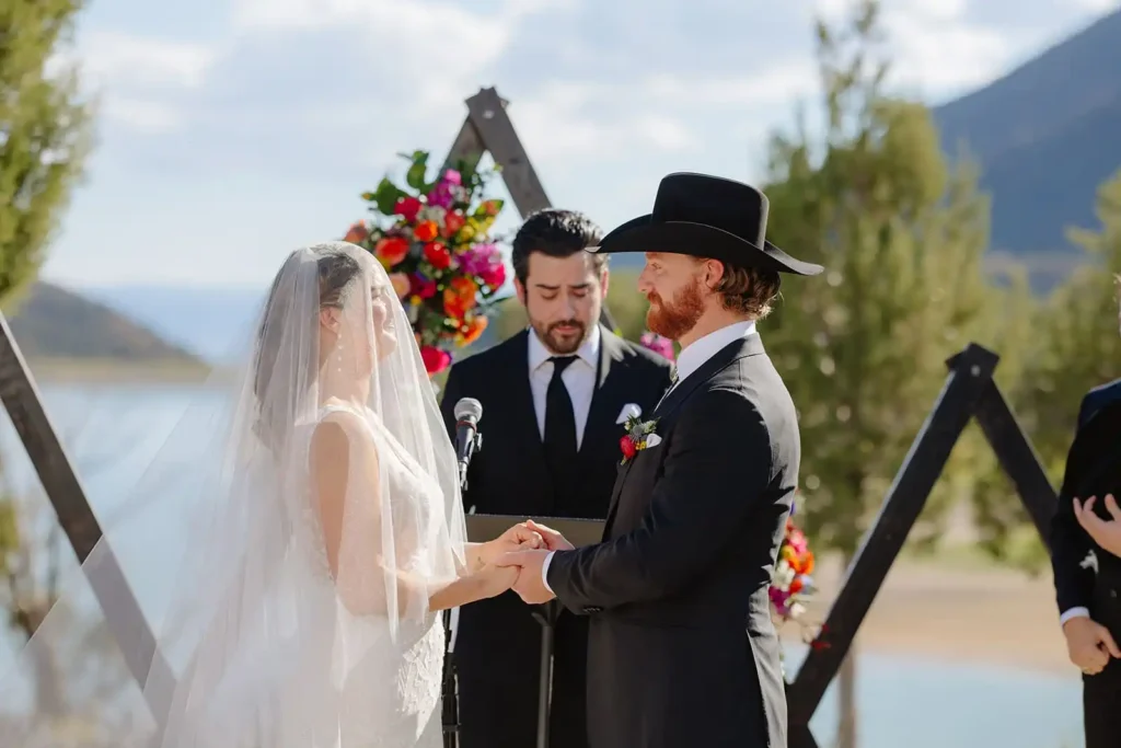 Couple holding hands during vows, framed by vibrant floral arch with lake behind them.