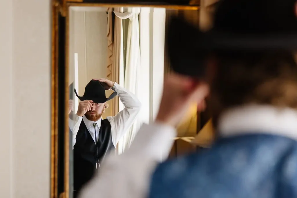 Editorial style photo of the groom putting on a cowboy hat in the mirror at Hotel Colorado on wedding day