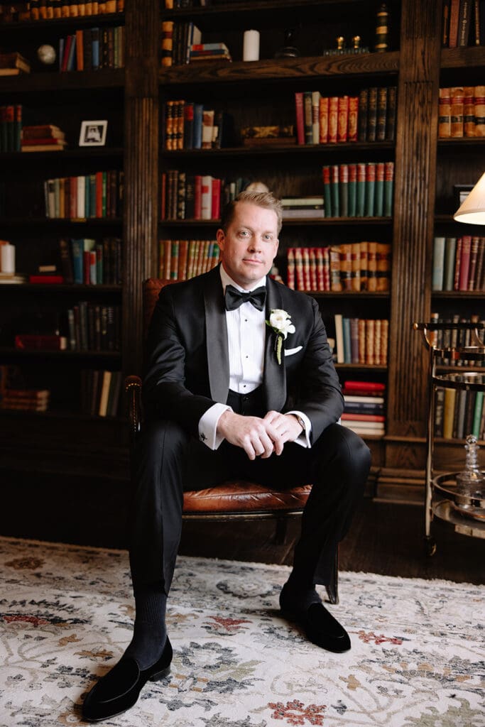 Manor House portraits in the library: A man in a tuxedo sits in a leather chair, exuding elegance. Behind him is a wall of bookshelves filled with colorful books, creating a sophisticated library ambiance.