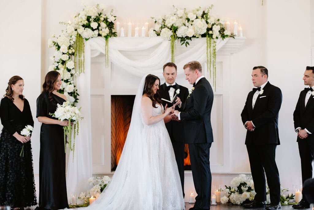 A bride and groom exchange rings at their winter wedding ceremony at The Manor House wedding venue, standing before a floral arch with attendants in black formal attire on either side.