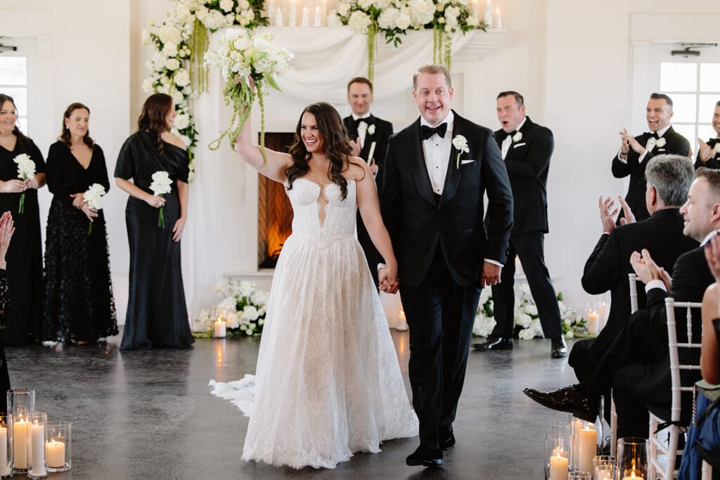 A bride and groom celebrating at their winter wedding ceremony at The Manor House wedding venue. They are standing in front of an elegant fireplace with attendants in black formal attire on either side.