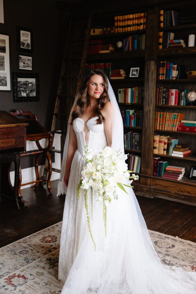 Manor House Wedding Venue portraits in the library: Bride in elegant white gown stands in a library, holding a large bouquet of white flowers. The room has dark wooden shelves filled with books, creating a classic, serene atmosphere.