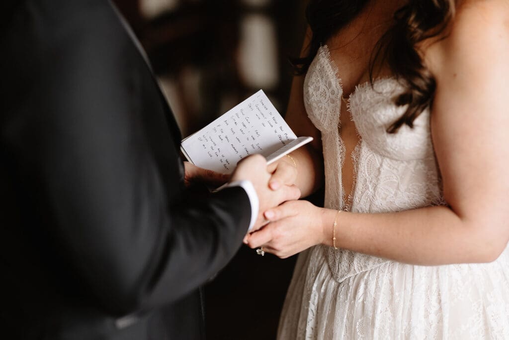 Intimate ceremony at a manor house wedding venue, bride in lace gown holding hands with groom as he reads handwritten vows from a notebook, romantic indoor wedding moment with warm, rustic elegance.