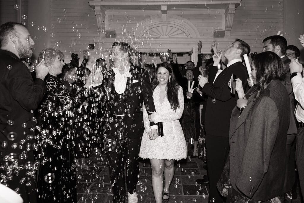 Black and white photo of a wedding exit at denver wedding venue, couple walking hand in hand through cheering guests blowing bubbles, joyful send-off moment with classic architectural backdrop near Denver.