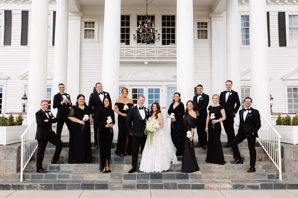 Elegant bridal party portrait at a manor house wedding venue, bride and groom centered on grand front steps with white columns, surrounded by bridesmaids in black dresses and groomsmen in tuxedos, classic estate wedding aesthetic with timeless architecture and formal group styling.