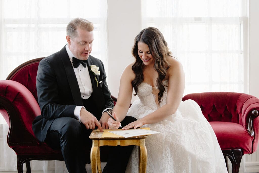 Bride and groom sign their marriage license in the bridal suite at the manor house on a red velvet couch during winter wedding